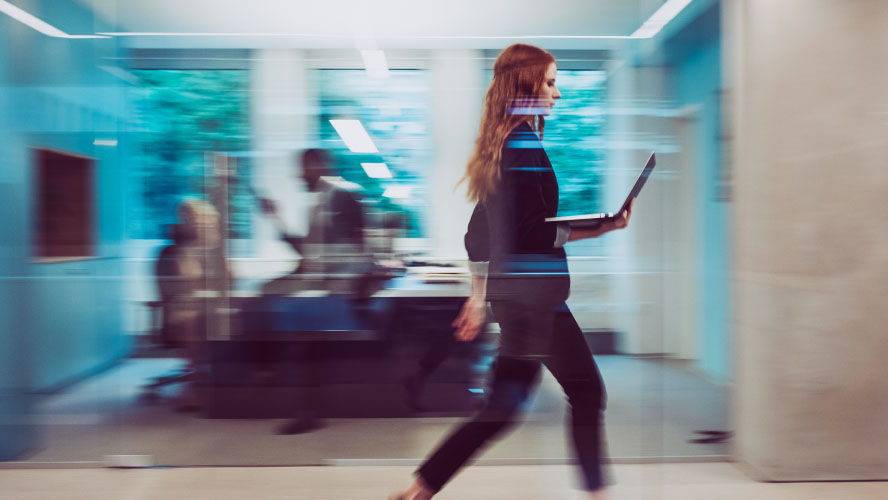 Une femme traverse avec assurance un immeuble de bureaux, munie d'un ordinateur portable, absorbée par son travail.