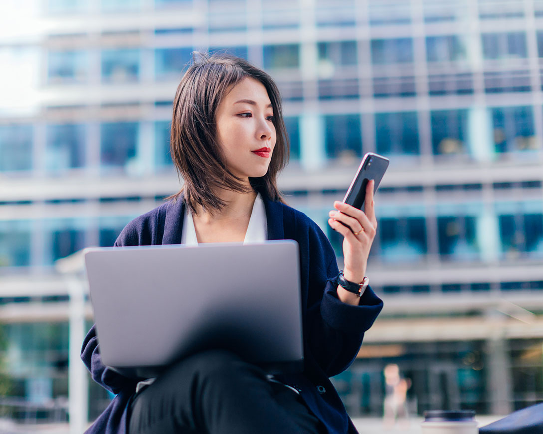 Optimization – A businesswoman working on a laptop and smartphone outside in the financial district